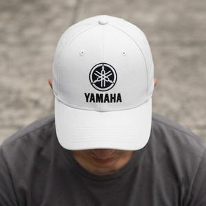 Könnte beinhalten: Eine weiße Baseballkappe mit dem schwarzen Yamaha-Logo und Schriftzug mittig auf der Vorderseite. Die Kappe verfügt über eine strukturierte Krone, einen gebogenen Schirm und gestickte Ösen zur Belüftung, was ein schlichtes Design ergibt.