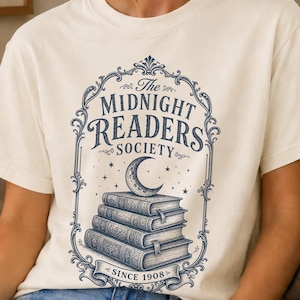 May include: An off-white short-sleeve t-shirt featuring a dark blue graphic print of a stack of books topped with a crescent moon inside an ornate frame. The text reads The Midnight Readers Society and Since 1908 in a decorative font.