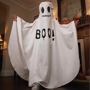 May include: A white ghost costume made of fabric, featuring a printed black face with two oval eyes and a stitched mouth. The word BOO! is printed in large, bold black letters across the centre of the chest. The garment drapes loosely over the body.