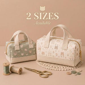 May include: Two rectangular fabric bags with top handles and gold zippers, featuring a bear and flower print. The smaller bag is sage green, the larger is cream. Includes wooden thread spools, metal scissors, and small buttons on the surface.