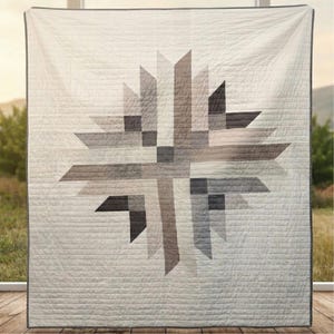 Puede incluir: Una colcha cuadrada con un patrón central de estrella compuesto por formas geométricas en tonos blanco, gris claro, carbón y marrón oscuro. La colcha tiene costuras horizontales en toda su superficie y un borde gris oscuro.