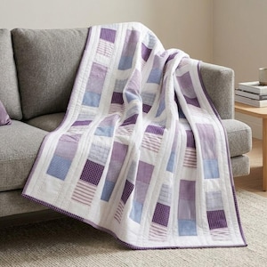 May include: A quilt featuring a patchwork design of vertical strips in shades of purple, lavender, and blue, alternating with solid white fabric panels. The pattern includes various textures such as gingham, stripes, and small checkered prints.