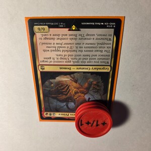 May include: A Magic: The Gathering trading card titled The Beast, featuring a 6/6 power and toughness rating, displayed alongside a round red plastic token with the text +1/+1 printed in black ink on its surface, oriented vertically.