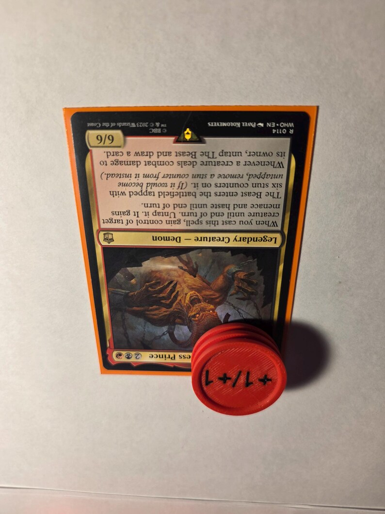 May include: A Magic: The Gathering trading card titled The Beast, featuring a 6/6 power and toughness rating, displayed alongside a round red plastic token with the text +1/+1 printed in black ink on its surface, oriented vertically.