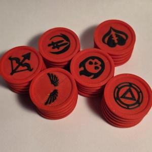 May include: A collection of red circular tokens stacked in small groups. Each token features a distinct black embossed symbol, including a bow and arrow, a pair of wings, a skull, a spade-like shape, and a triangle enclosed within a circular frame.