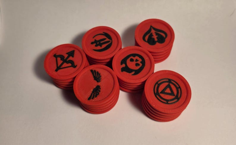 May include: A collection of red circular tokens stacked in small groups. Each token features a distinct black embossed symbol, including a bow and arrow, a pair of wings, a skull, a spade-like shape, and a triangle enclosed within a circular frame.