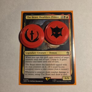 May include: A Magic: The Gathering trading card titled The Beast, Deathless Prince, featuring a legendary demon creature. Two round, bright orange plastic tokens with black embossed symbols, one a sword and one a mask, rest on the card surface.