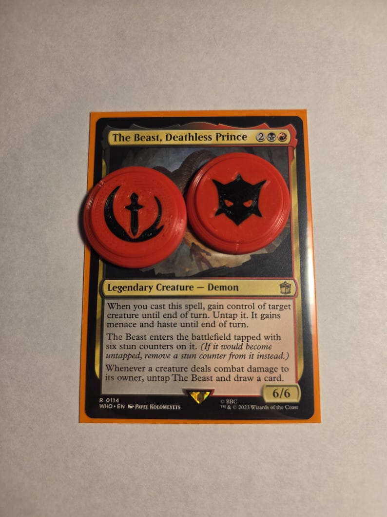 May include: A Magic: The Gathering trading card titled The Beast, Deathless Prince, featuring a legendary demon creature. Two round, bright orange plastic tokens with black embossed symbols, one a sword and one a mask, rest on the card surface.