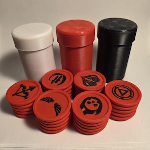 Puede incluir: Tres recipientes cilíndricos en blanco, rojo y negro se sitúan detrás de varias pilas de fichas circulares rojas. Cada ficha presenta un símbolo negro en relieve, incluyendo una flecha, alas, una calavera y varios diseños geométricos.