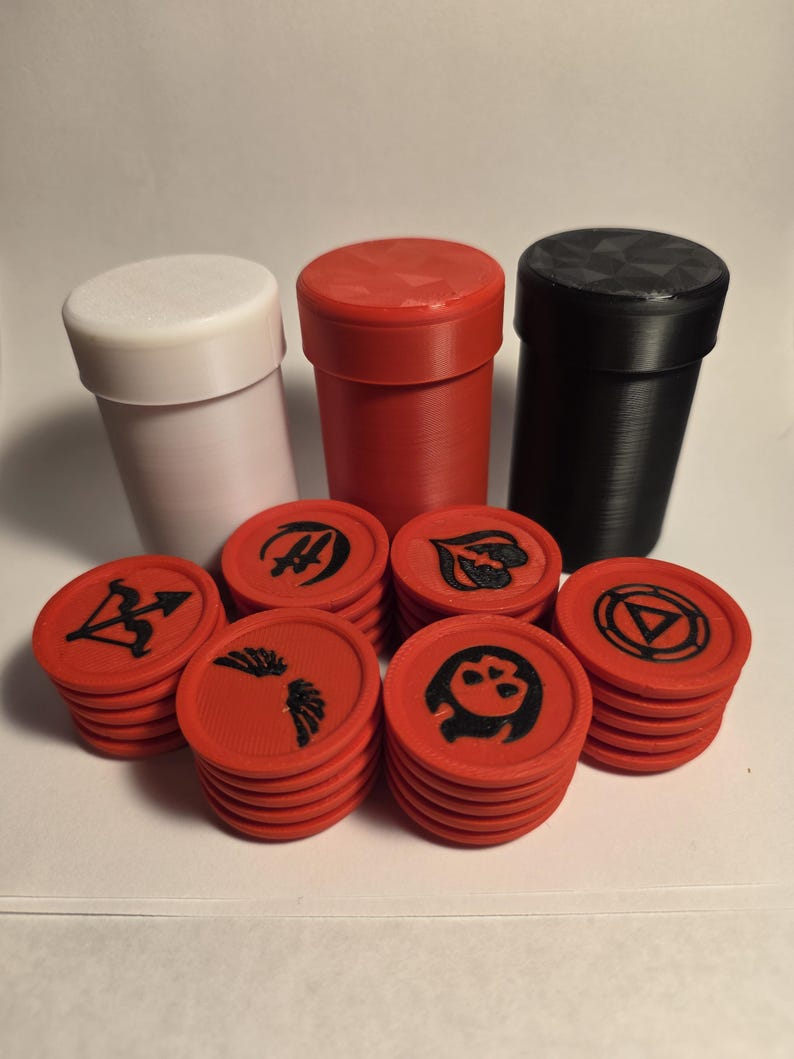 May include: Three cylindrical containers in white, red, and black stand behind several stacks of red circular tokens. Each token features a distinct black embossed symbol, including an arrow, wings, a skull, and various geometric designs.
