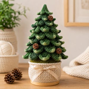 May include: A crocheted Christmas tree with layered green foliage and small brown pinecone accents. The tree is set in a beige crocheted pot decorated with a smooth, light-colored ribbon tied in a bow at the center, resting on a wooden base.