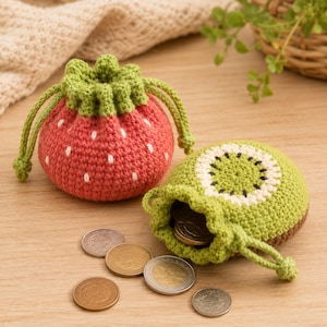 Puede incluir: Dos pequeñas bolsas redondas de ganchillo con cordón. Una bolsa es roja con semillas blancas bordadas, parecida a una fresa. La otra es verde con un centro blanco y negro, parecida a un kiwi. Varias monedas de metal se encuentran cerca.