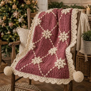 Puede incluir: Una manta de ganchillo rectangular en color rosa empolvado oscuro con un patrón de rombos en color crema y motivos de copos de nieve en cada intersección. La manta tiene un borde festoneado en crema y grandes pompones crema en las esquinas.