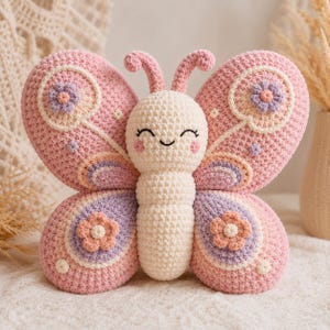 May include: A crocheted butterfly plush toy featuring a cream-colored body and large pink wings. The wings are decorated with lavender and peach-colored crocheted flower appliques, small white dots, and a smiling face with pink cheeks.