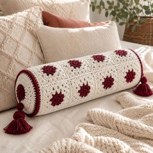 May include: A cylindrical bolster pillow featuring a cream-colored crochet granny square pattern with dark red floral centers. The ends of the pillow are finished with dark red crochet borders and matching dark red decorative yarn tassels.
