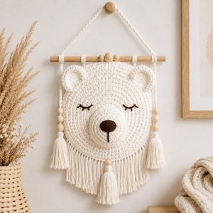 Op de afbeelding: Een witte gehaakte berenwandhanger met een rond gezicht, gesloten ogen en een donkerbruine neus. Het item hangt aan een lichte houten stok met een bijpassend koord, versierd met houten kralen en lange crèmekleurige kwastjes.