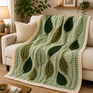 May include: A crochet blanket featuring a textured pattern of raised leaves in various shades of green, including sage, olive, and forest green. The blanket has a cream-colored border and is draped over a sofa, showcasing intricate stitch work.