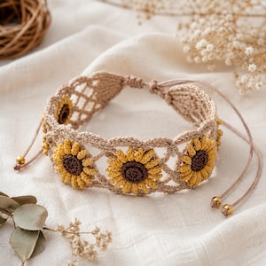 Op de afbeelding: Een beige macramé armband met drie gehaakte zonnebloemmotieven met gele bloemblaadjes en donkerbruine harten. De band is gemaakt met een open rasterpatroon en bevat verstelbare dunne koorden afgewerkt met kleine metalen gouden kralen.