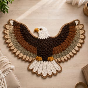 May include: A crochet rug shaped like a bald eagle with wings spread wide. The eagle features a white head and tail, a dark brown body, and wings layered with shades of brown, tan, and cream. It has a yellow beak and yellow talons.