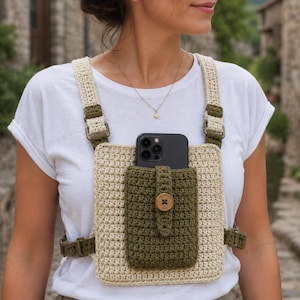 May include: A cream and olive green crochet chest bag featuring a rectangular base, a central olive green pocket with a button closure, and adjustable straps with metal hardware. A black smartphone is tucked into the front pocket of the bag.