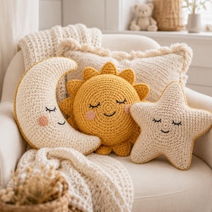 May include: A set of three crochet cushions featuring a cream crescent moon, a yellow sun, and a cream star. Each cushion has embroidered closed eyes, a small smile, and pink circular cheeks. The cushions are textured with visible yarn stitches.