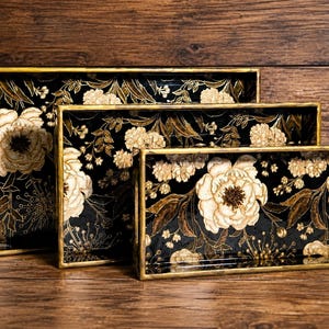 May include: A set of three rectangular nesting trays featuring a black base with a gold floral pattern. Each tray has a metallic gold rim and integrated side handles, displaying a consistent botanical design of large peonies and delicate foliage.