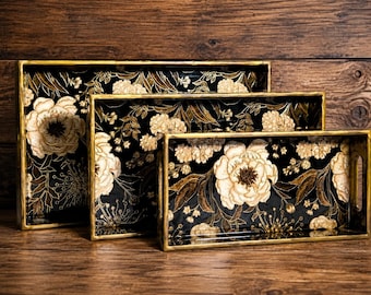 May include: A set of three rectangular nesting trays featuring a black base with a gold floral pattern. Each tray has a metallic gold rim and integrated side handles, displaying a consistent botanical design of large peonies and delicate foliage.