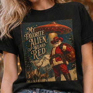 Pode incluir: Uma t-shirt preta de manga curta com uma estampa gráfica do Pai Natal num cenário nevado com renas e um disco voador por cima. O texto diz "MY FAVORITE ALIEN WEARS RED". O design apresenta uma estética vintage com aspeto desgastado.