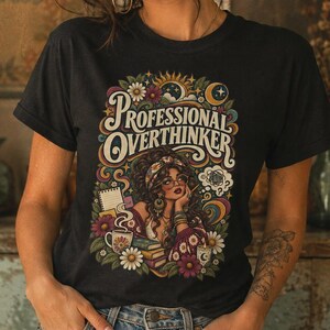 Pode incluir: Uma t-shirt preta com uma estampa gráfica colorida com o texto "PROFESSIONAL OVERTHINKER" acima de uma ilustração de uma pessoa rodeada por flores, livros, uma caneca de café, um bloco de notas e símbolos celestiais como sol e lua.