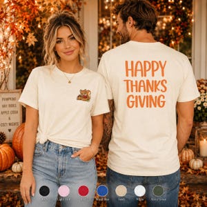 Puede incluir: Una camiseta de manga corta color arena con un pequeño gráfico de una ardilla sosteniendo una cesta en el pecho izquierdo. La parte posterior de la camiseta muestra el texto naranja HAPPY THANKS GIVING en una fuente sans-serif centrada.