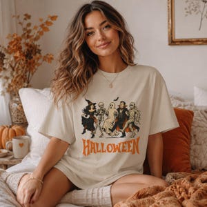 May include: A cream-colored short-sleeve t-shirt featuring a graphic print of a witch, skeleton, ghost, vampire, and mummy dancing together. Below the characters, the word HALLOWEEN is printed in large, bold, orange, distressed-style lettering.
