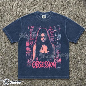 May include: A dark blue short-sleeve t-shirt featuring a pink graphic print of a person with long hair. The text reads I LOVE YOU SO SO SO SO MUCH, BE CAREFUL WHO YOU WISH FOR, YOU WISHED FOR THIS, and OBSESSION, along with heart icons.