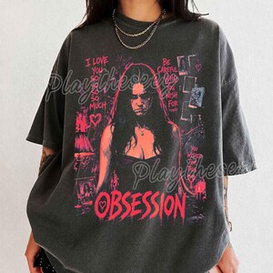 May include: A charcoal gray short-sleeve t-shirt featuring a graphic print of a person with long dark hair. The design includes pink text that reads I LOVE YOU SO SO SO MUCH, BE CAREFUL WHO YOU WISH FOR, YOU WISHED FOR THIS, and OBSESSION.