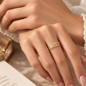 Könnte beinhalten: Ein goldfarbener Ring mit dem Namen Sara in einer verzierten, gotischen Schriftart. Der Ring ist schmal und poliert und zeigt den individuellen Namen deutlich auf dem Finger. Das Design ist schlicht und fokussiert auf die Schrift.