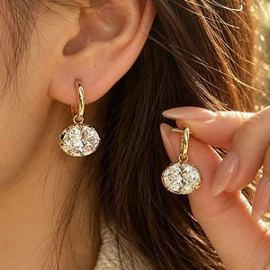 May include: A pair of gold hoop earrings featuring oval-shaped clear faceted stones suspended from the hoops. The earrings have a polished metallic finish and the stones are set in a secure prong setting, reflecting light with a brilliant shine.
