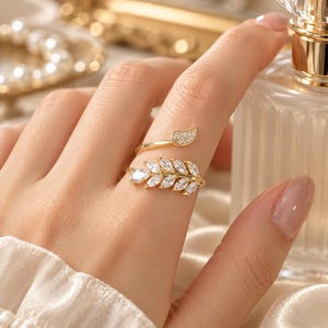 Puede incluir: Un anillo abierto de tono dorado con un diseño en forma de hoja adornado con piedras transparentes de corte marquesa y un acento de hoja más pequeño incrustado con diminutos cristales pavé. La banda es ajustable y envuelve el dedo con un acabado metálico pulido.