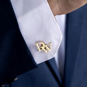 Może przedstawiać: Złoty metalowy spinka do mankietów z literami DW w stylizowanej, połączonej czcionce. Spinka jest przymocowana do białego materiałowego mankietu koszuli z długim rękawem, noszonej pod ciemnogranatową marynarką typu blazer.