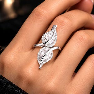 May include: A silver-toned bypass ring featuring two leaf-shaped motifs. Each leaf is encrusted with small, clear, rectangular baguette-cut stones in the center, surrounded by a border of tiny, round, clear pavé-set stones along the edges.