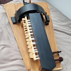 Könnte beinhalten: Eine Drehleier mit einem Korpus aus hellem Naturholz, einem schwarzen zentralen Wirbelkasten und einer Reihe von hellen Holztasten. Das Instrument verfügt über eine Metallkurbel an der Oberseite, dunkelbraune Wirbel und sichtbare Saiten.