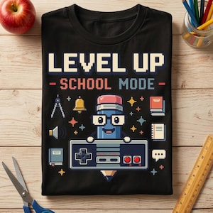 May include: A black t-shirt featuring a pixel art graphic with the text "LEVEL UP - SCHOOL MODE". The design includes a blue pencil character wearing glasses and holding a video game controller, surrounded by school icons like a bell and books.
