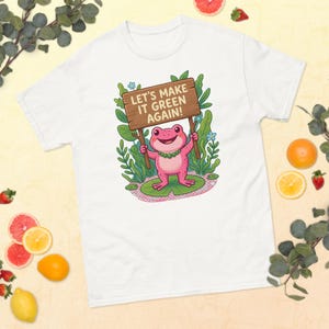 May include: A white t-shirt featuring a graphic of a pink frog standing on a lily pad, holding a wooden sign that reads "LET'S MAKE IT GREEN AGAIN!" The frog is surrounded by green foliage and small blue flowers, with a beaded necklace around its neck.