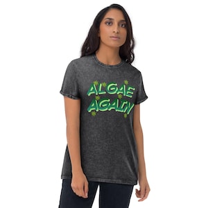 Könnte beinhalten: Ein anthrazitfarbenes T-Shirt in Acid-Wash-Optik mit dem Text ALGAE AGAIN in einer geschichteten grünen Schriftart. Das Design enthält kleine grüne sternförmige Symbole, die um den Schriftzug herum verteilt sind, der mittig auf der Brust des kurzärmeligen Kleidungsstücks platziert ist.