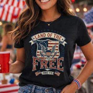 Könnte beinhalten: Ein schwarzes Kurzarm-T-Shirt mit einem abgenutzten Grafikdruck, der einen Schild, einen Adler sowie den Text "LAND OF THE FREE" und "SINCE 1776" zeigt. Das Design enthält rote, weiße und blaue Farben mit Sternen und kleinen Flaggen.