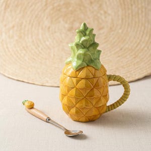May include: A ceramic pineapple-shaped mug with a removable green lid and a textured yellow body. The mug features a twisted green handle and comes with a matching metal spoon that has a small wooden handle topped with a miniature pineapple charm.