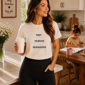 May include: A cream-colored short-sleeve t-shirt featuring the black text "TINY HUMAN MANAGER" printed on the front. The shirt has a crew neckline and is paired with solid black leggings that include side pockets on the upper thigh area.