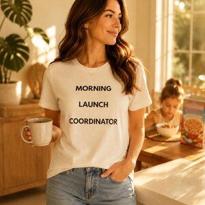 May include: A white short-sleeved t-shirt featuring the black text MORNING LAUNCH COORDINATOR printed on the front. The shirt is paired with blue denim jeans and is worn while holding a light-coloured ceramic mug in a bright, sunlit indoor setting.