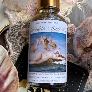 Könnte beinhalten: Eine Glasflasche mit goldenem Tropfverschluss, die Öl enthält. Das Etikett zeigt den Text THE WITCHING HOUR Aphrodite Spell Oil, eine Abbildung einer liegenden Figur mit Putten sowie den Text A DEVOTIONAL OIL FOR CONNECTING WITH APHRODITE, EMBODY THE DIVINE FEMININE ENERGY AND EVOKE LOVE AND BEAUTY WITHIN YOUR LIFE.