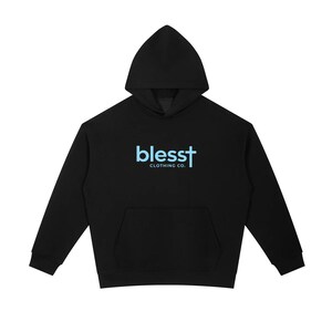 May include: A black long-sleeve hooded sweatshirt featuring a front kangaroo pocket. The center of the chest displays the light blue text "blesst" with a cross replacing the letter "t", followed by the white text "CLOTHING CO." underneath.