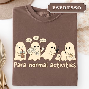 May include: A brown t-shirt featuring a graphic of five cream-colored ghosts. The ghosts are holding items like a drink, documents, a stopwatch, a walkie-talkie, and a tablet. The text "d-a-t-a" appears above them, and "Para normal activities" is below.