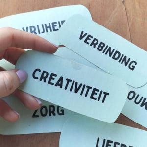 Op de afbeelding: Een verzameling lichtgroene rechthoekige kaartjes met afgeronde hoeken, elk voorzien van een enkel woord in een vetgedrukt zwart schreefloos lettertype. De zichtbare woorden zijn CREATIVITEIT, VERBINDING, ZORG, LIEFDE en VRIJHEID.